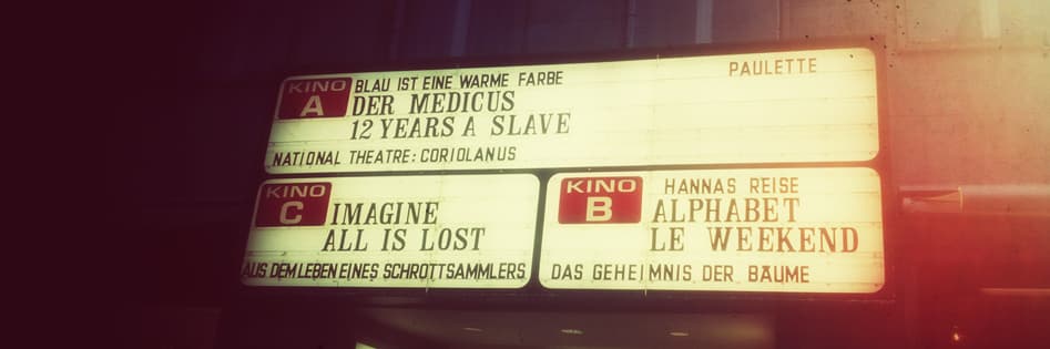 Backlit cinema marquee with red “Kino A”, “Kino B” and “Kino C” panels listing films such as “Der Medicus” and “12 Years a Slave”, in warm vintage tones